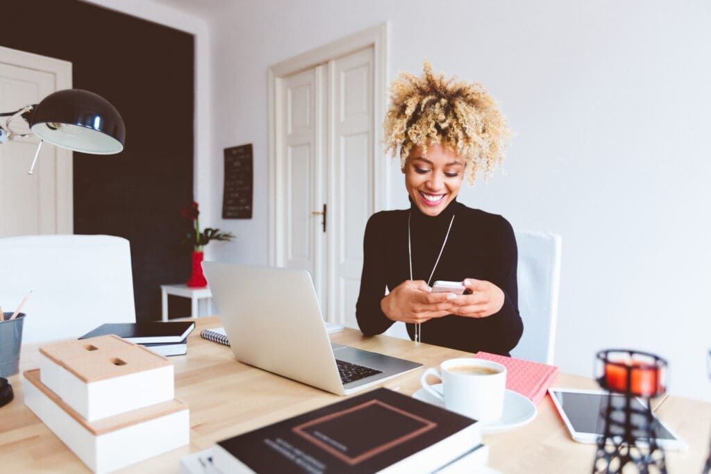 Young Afro American woman sits at her desk in her home office with her laptop open, smiling and using her smartphone.