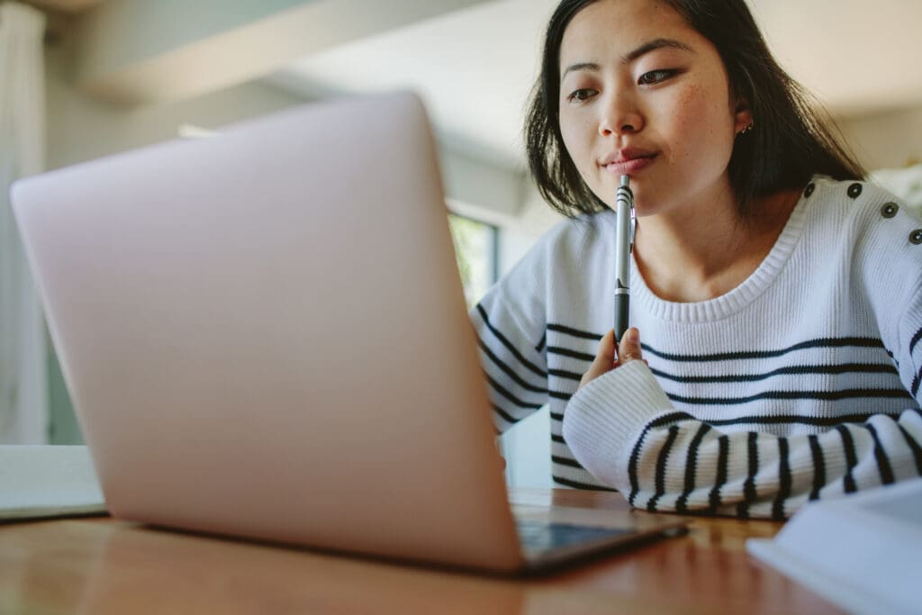 Young Asian woman sits at a table at home, holding a pen, focused on the laptop in front of her while she takes a virtual campus tour.